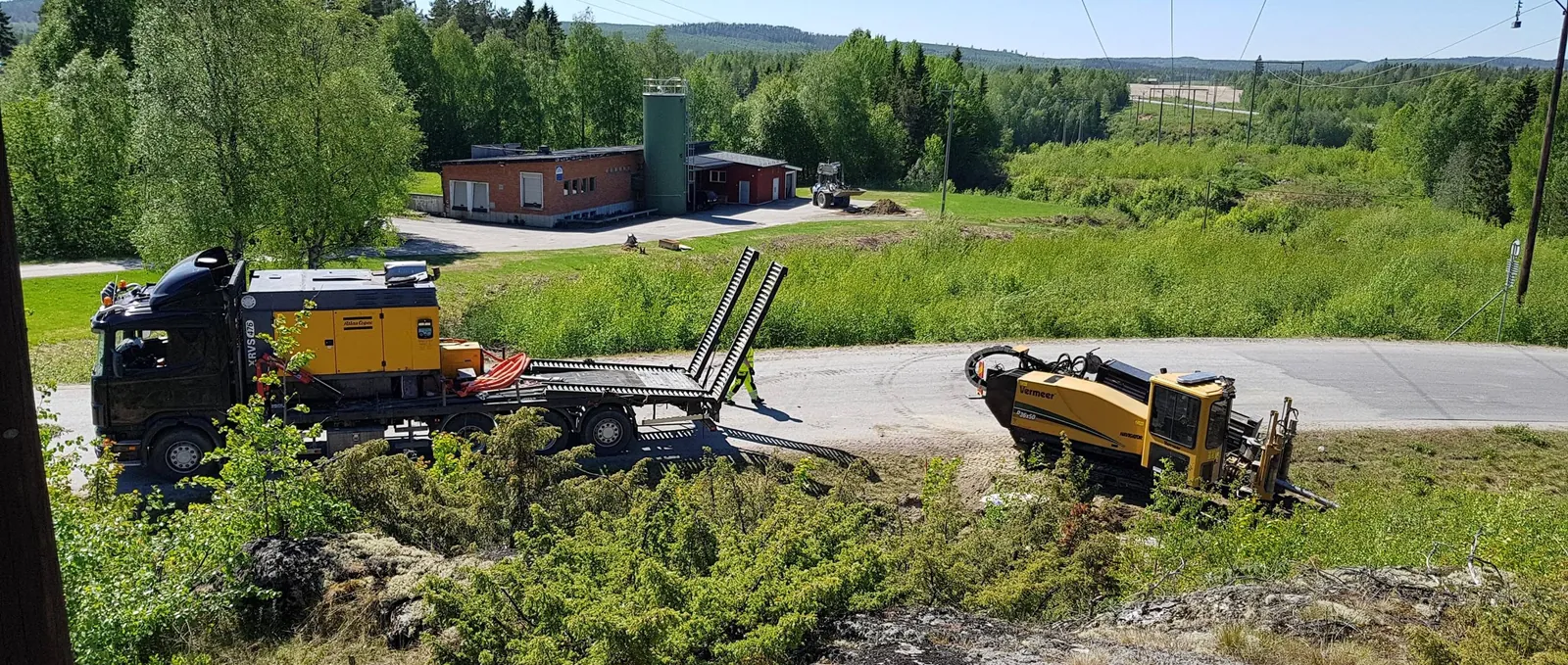 Gul borrigg för styrd borrning etablerad vid en asfaltväg i skogsmark. Till vänster en lastbil med kompressor på flaket.